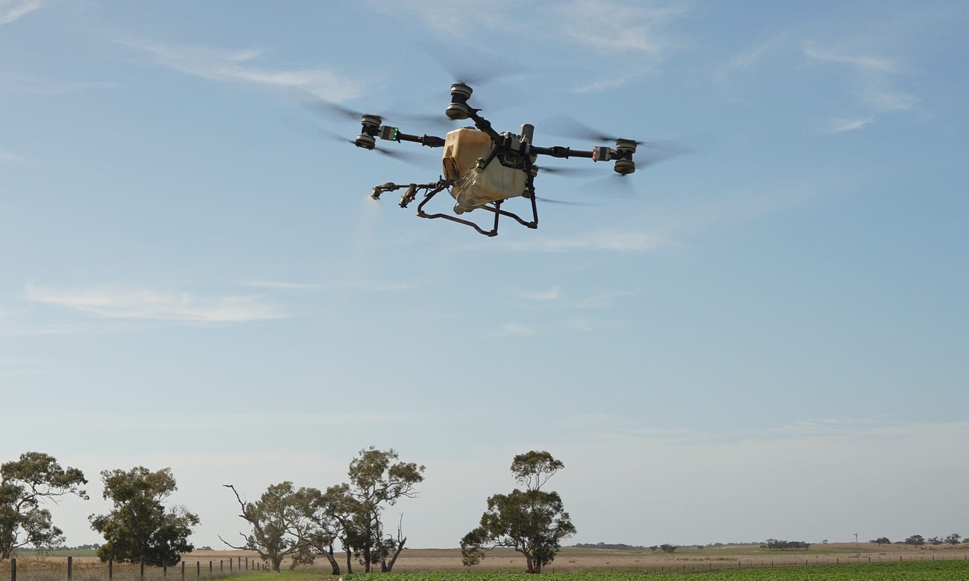 An AGX spray drone hovering over a paddock track with gum trees along the fence line